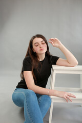 stylish photo shoot beautiful model with black hair. girl in jeans and black turtleneck on a gray background. girl sitting on a white chair and posing for a photo