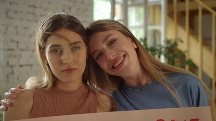 Looking to each other, two handsome young girls are standing in the studio and showing a lgbt poster at the camera