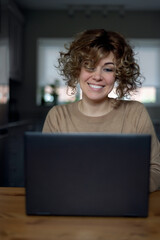 Portrait of a cheerful woman who is looking at laptop in her kitchen, working from home. Using computer for video call, looking at screen or reading news