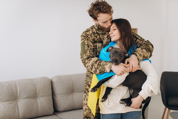 A Ukrainian couple, a soldier in military uniform and a girl wrapped in a Ukrainian flag hold a dog in their arms, happy together.