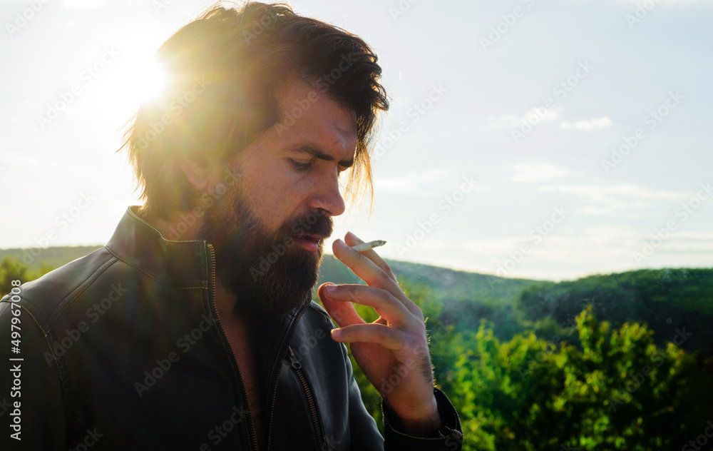 Wall mural Handsome hipster with beard in leather jacket smoking cigarette or tobacco. Serious bearded man.