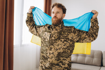 Portrait of an emotional young bearded Ukrainian patriot soldier in military uniform holding a flag in the office