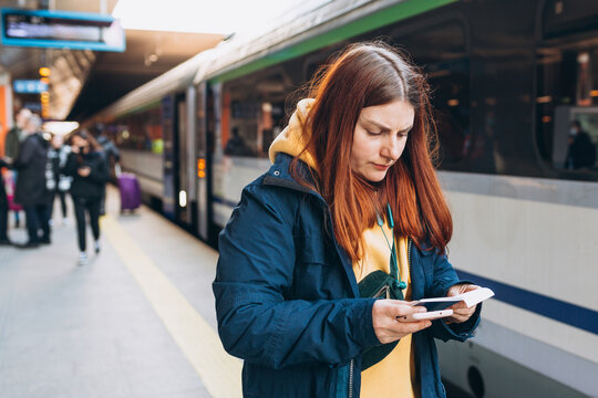 Young redhhead woman with backpack checking her ticket and using smartphone on background train. Railroad transport concept. Girl standing near a platform with blurred railway station.