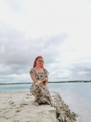 A beautiful girl with pink hair walks near the water on the beach. Honeymoon, early pregnancy woman. Emerald water, vacation, family day off. Girl in a boho style dress on the background of nature