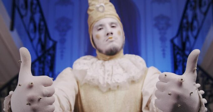 A make-up cool dramatic funny mime guy on a blue theatrical stage shows thumbs up gesture. A theater actor dressed as an old Baroque harlequin. Copy space theme for promo video