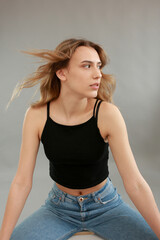 Beautiful girl model appearance in light jeans and black T-shirt on a gray studio background. Girl model sitting on a white chair. Beautiful blonde shows poses for a photo shoot