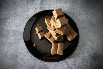 Pieces of different milk chocolate on black dish on gray table, view from above