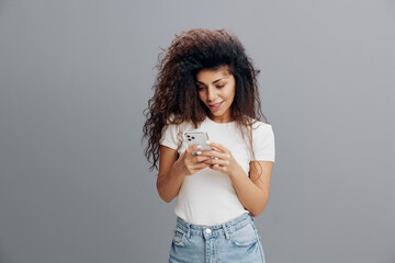 Smiling curly Latin tanned woman in casual white t-shirt posing isolated on grey wall background studio portrait. People lifestyle concept. Mock up copy space. Using mobile phone, typing sms message
