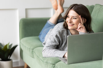 Young beautiful smiling woman using laptop lying on the couch. Concept of freelance job, home office. Female using technology for learning, shopping, e-commerce, communication, social media