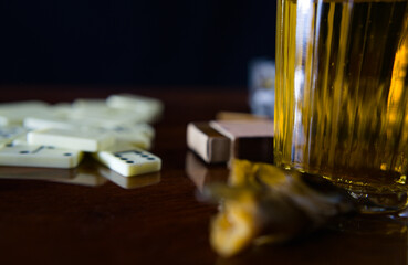 A glass of light beer in close-up with a piece of salted fish, scattered dominoes and a matchbox in the background. Still life - spending leisure time playing dominoes. - Powered by Adobe