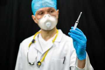 A doctor in a white coat and a syringe in his hand. A man in a medical mask and a medical gown on a black background. Vaccination against coronavirus and covid19