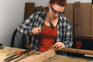 Young red-haired carpenter hammering a nail into a wooden board in her small carpentry workshop.