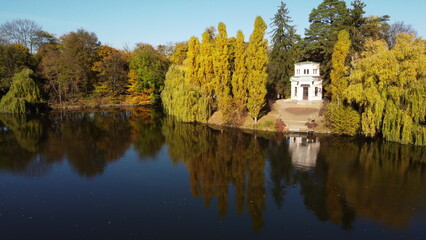 Fototapeta na wymiar Aerial Drone View Flight Over water mirror surface of lake in park on windless sunny autumn day. Trees with yellow leaves on banks. In middle of lake - white motor boat. White building on the coast