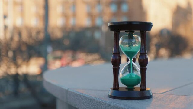 An hourglass made of wood and glass with greenish sand granules stands on the city parapet in the center of the metropolis. Life is boiling in the background. Cars are passing in the out-of-focus.