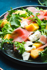 Delicious summer fresh salad with cantaloupe melon, prosciutto, soft cheese and arugula on blue table background, top view, copy space