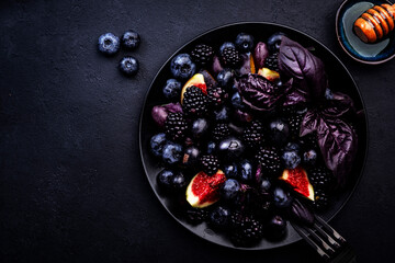 Summer blue and black berries fruit vegan salad: blueberries, blackberries, grapes, figs and purple basil with honey dressing on dark background, top view