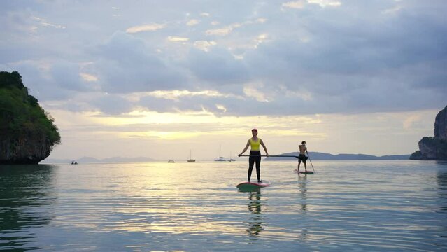 4K Confidence Asian man and woman standing on paddle board paddleboarding in the sea together at summer sunset. Healthy male and female enjoy outdoor lifestyle and water sports on holiday vacation
