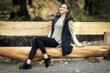 Portrait of a slender joyful young woman in full growth sitting on a wooden bench in a park outdoors