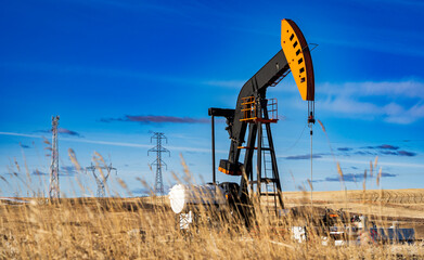 An oil pump jack working on an agriculture field with oil and gas equipment and distant power lines in Rocky View County Alberta Canada.