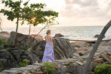 Woman in purple dress walk on a stone walkway at the beach, enjoy romantic sunset moment.