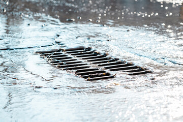 The grate of the storm sewer after the rain. The water drains into the storm drain. Sun glare, defocused background - obrazy, fototapety, plakaty
