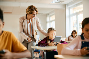 Senior teacher assists schoolgirl in using digital tablet during a class at school.