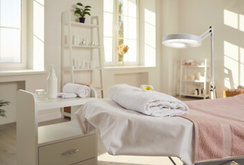 Beautiful modern interior of new spa salon, beautician's office, beauty parlor, or massage room with magnifier lamp, bed covered with white towels, and shelf with candle and facial skin care products