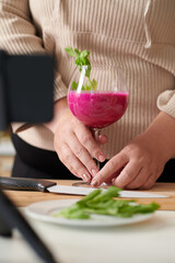 Hands of woman putting glass of delicious red smoothie with arugula she made for breakfast on table in front of smartphone