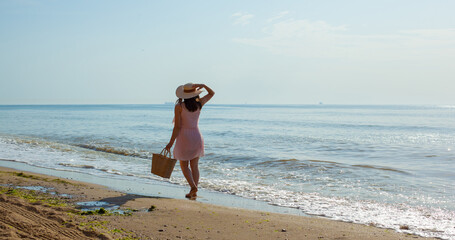 Happy young girl in a pink dress, straw hat and with summer bag going on the waves of the sea at noon. Relaxing unforgettable vacation at sea in summer. Enjoying summertime. Playing in water.