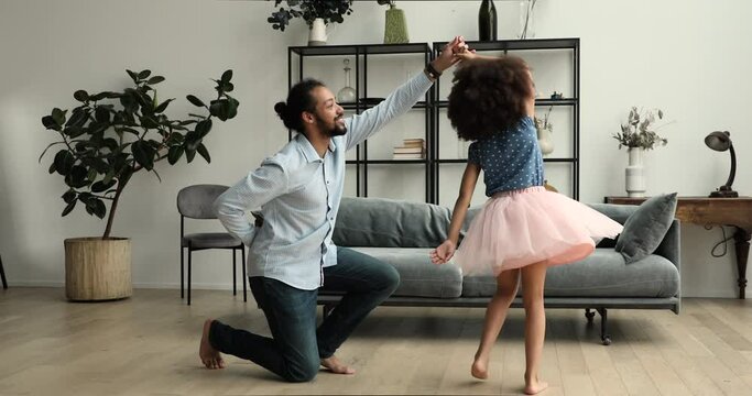 Caring Black dad stand on one knee hold hand of preteen daughter wearing fluffy skirt teach excited child to dance waltz. Father tween girl play princess knight dance on imaginary ball at living room