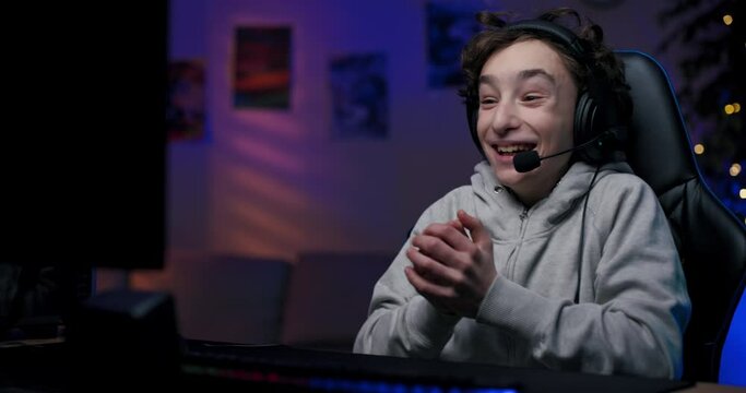 Pro gamer spends time in front of monitor playing computer games, boy claps hands wins round, has headset through which he talks while joking with team room lit with neon lights.