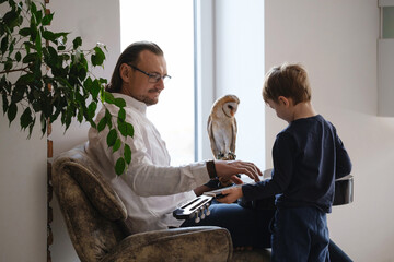 Man with owl and son at home playing guitar. Unusual pets and human animal friendship relationships. Wild bird on hand glove tenderness and love. Family leisure activities