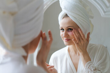 Skin care. Woman applying patches under her eyes, taking care her facial skin standing near mirror in at home. Young woman enjoying skin care using modern beauty products in robe and towel on her head