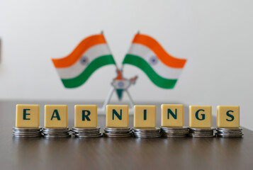 Letter blocks arranged as 'EARNINGS' on pile of coins and blur Indian flag in the background.  Main...