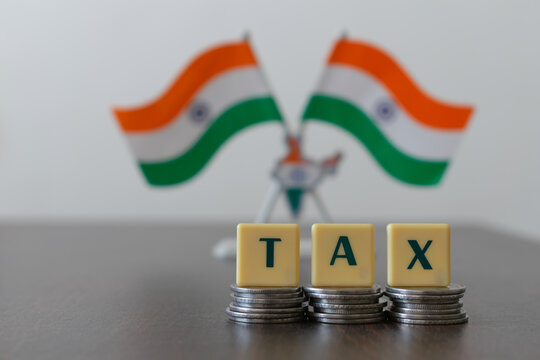 Letter blocks arranged as 'TAX' on pile of coins and blur Indian flag Tricolor in the background.  Main focus is on letter blocks and coins.