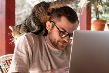 Cat sitting on the young adult man in glasses while man using his laptop working remotely.