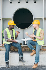 Content young multi-ethnic construction coworkers sitting on concrete beam with steel bars and chatting while drinking coffee during break