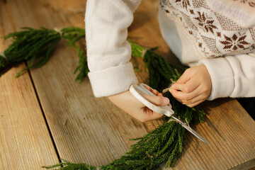 girl making cedar wreath on wooden craft table. child cutting a sisal thread.