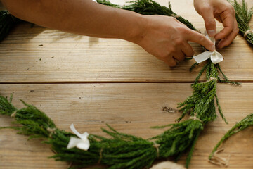 person holding white ribbon bows on cedar wreath on wooden craft table. space for copy.