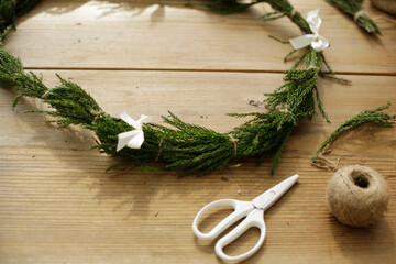 scissors, sisal twine and cedar leaves wreath tied up with white ribbon bows on wooden craft table. crafting outdoor. space for copy.