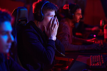 Shocked teenage gamer in headphones covering mouth and looking at monitor while losing game at...