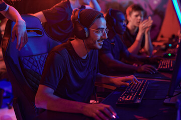 Smiling young professional gamer in hat and eyeglasses using computer while participating in esports tournament © Mediaphotos