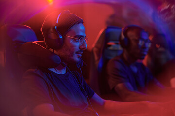 Concentrated young mixed race gamer in headset and glasses sitting in red light and playing online game - obrazy, fototapety, plakaty