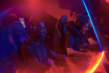 Multiracial team of young professional gamers in headsets sitting in row and playing online game in esports bar