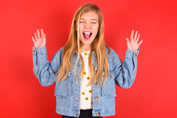 Emotive blonde little kid girl wearing denim jacket over red background laughs loudly, hears funny...