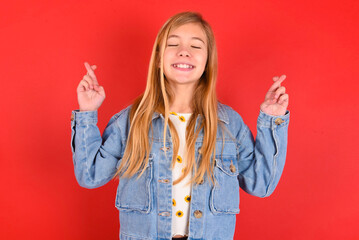 blonde little kid girl wearing denim jacket over red background has big hope, crosses fingers, believes in good fortune, smiles broadly. People and wish concept