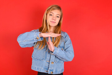 blonde little kid girl wearing denim jacket over red background being upset showing a timeout gesture, needs stop, asks time for rest after hard work, demonstrates break hand sign