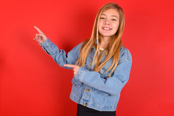 blonde little kid girl wearing denim jacket over red background indicating finger empty space showing best low prices, looking at the camera
