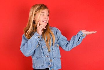 Funny blonde little kid girl wearing denim jacket over red background holding open palm new product. I wanna buy it!