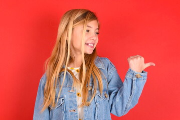 Charming blonde little kid girl wearing denim jacket over red background looking at copy space having advertisements